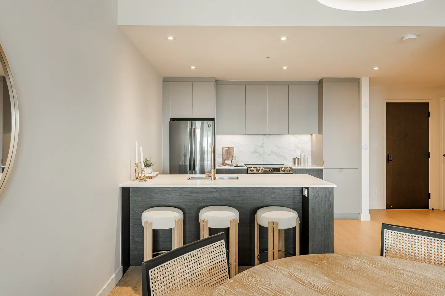 Modern kitchen with gray cabinets, stainless steel fridge, and island with white stools. A round wooden table and chairs are in the foreground.