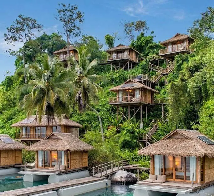 Luxury resort featuring thatched-roof bungalows on stilts and floating platforms amidst lush green tropical foliage.