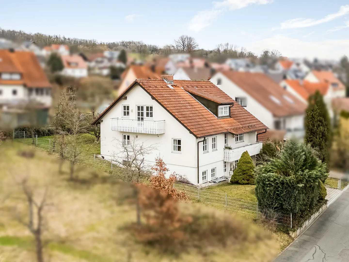 Two-story white house with a red tile roof and white balconies, set in a green yard with trees and a blurred background of other houses.