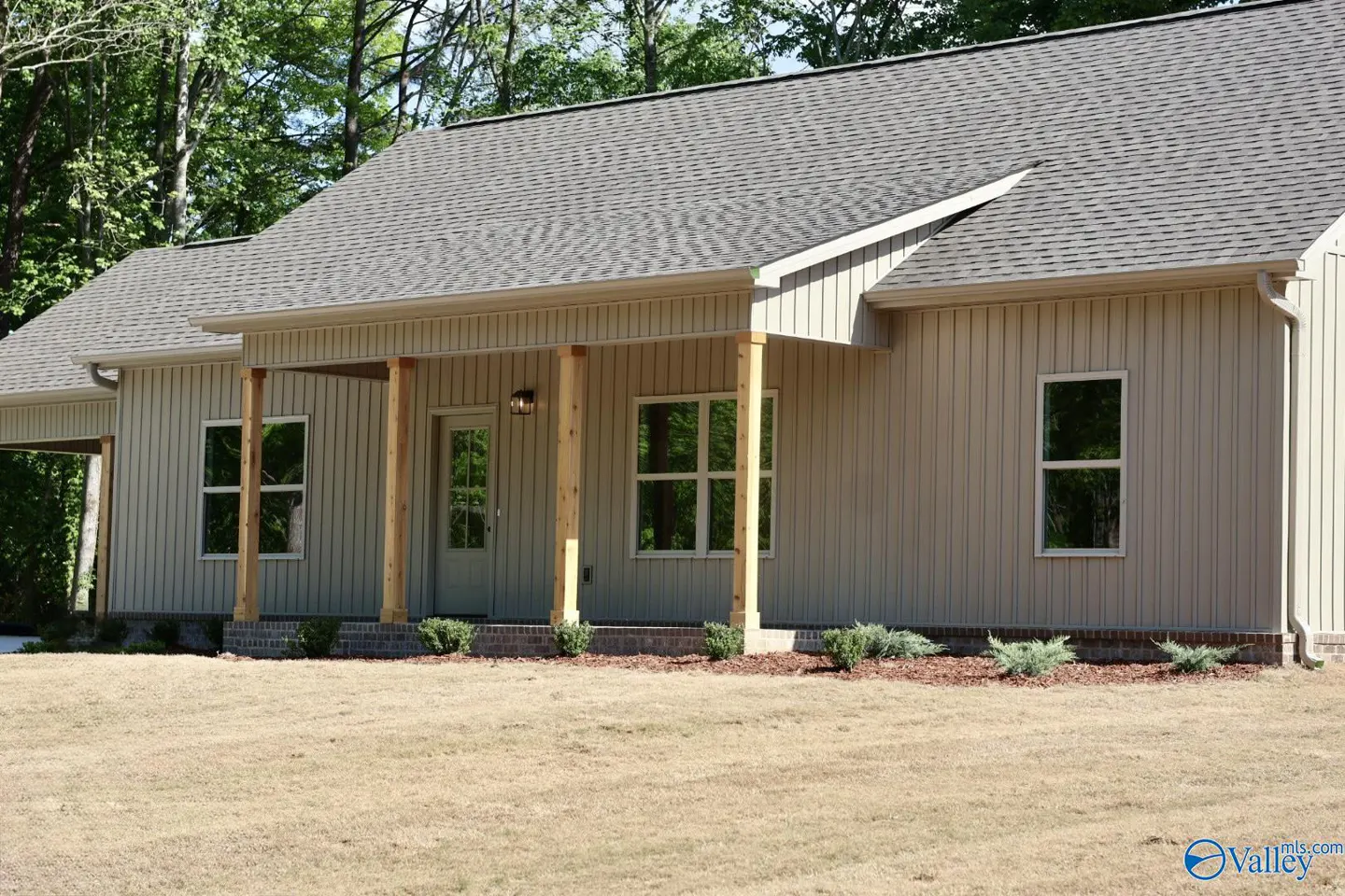 Tan house with gray roof and wooden porch columns. Green trees in background, brown grass in foreground.