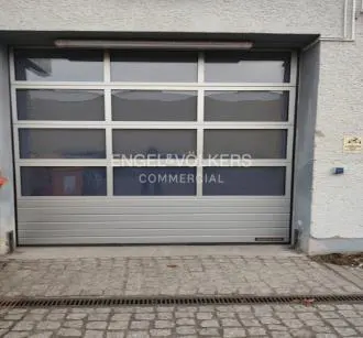 Silver garage door with glass panels and "Engel & Völkers Commercial" logo. Cobblestone pavement in front.