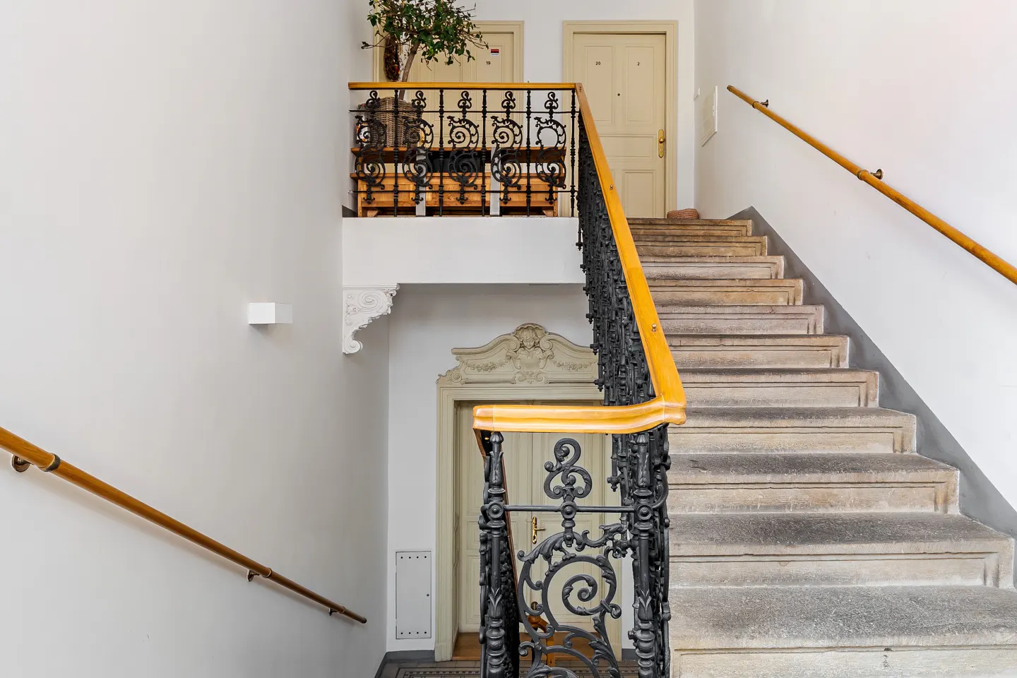 Interior view of a staircase with stone steps, wooden handrails, and ornate black iron railings in a white-walled building.