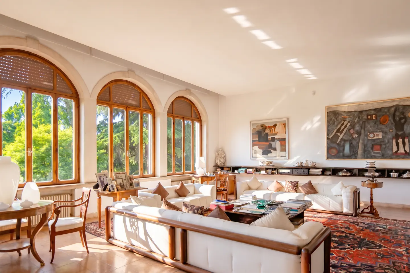 Bright living room with white sofas, wood trim windows, and abstract art. Sunlight streams in, illuminating the red patterned rug.