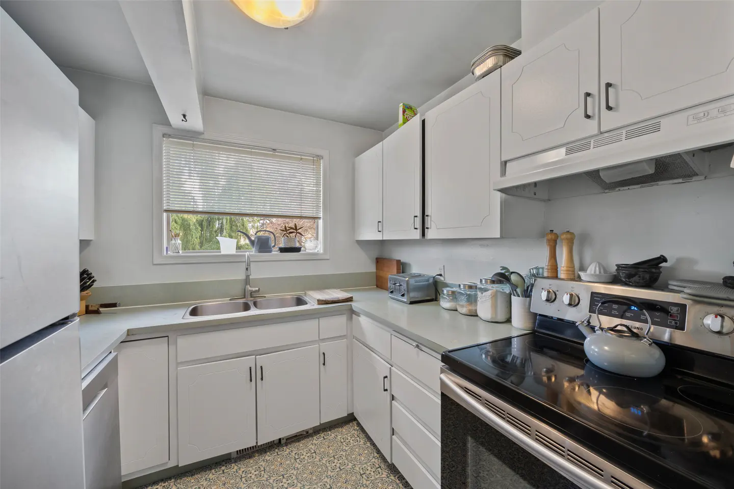 Bright kitchen with white cabinets, stainless steel appliances, and a window above the sink. A kettle sits on the black stovetop.