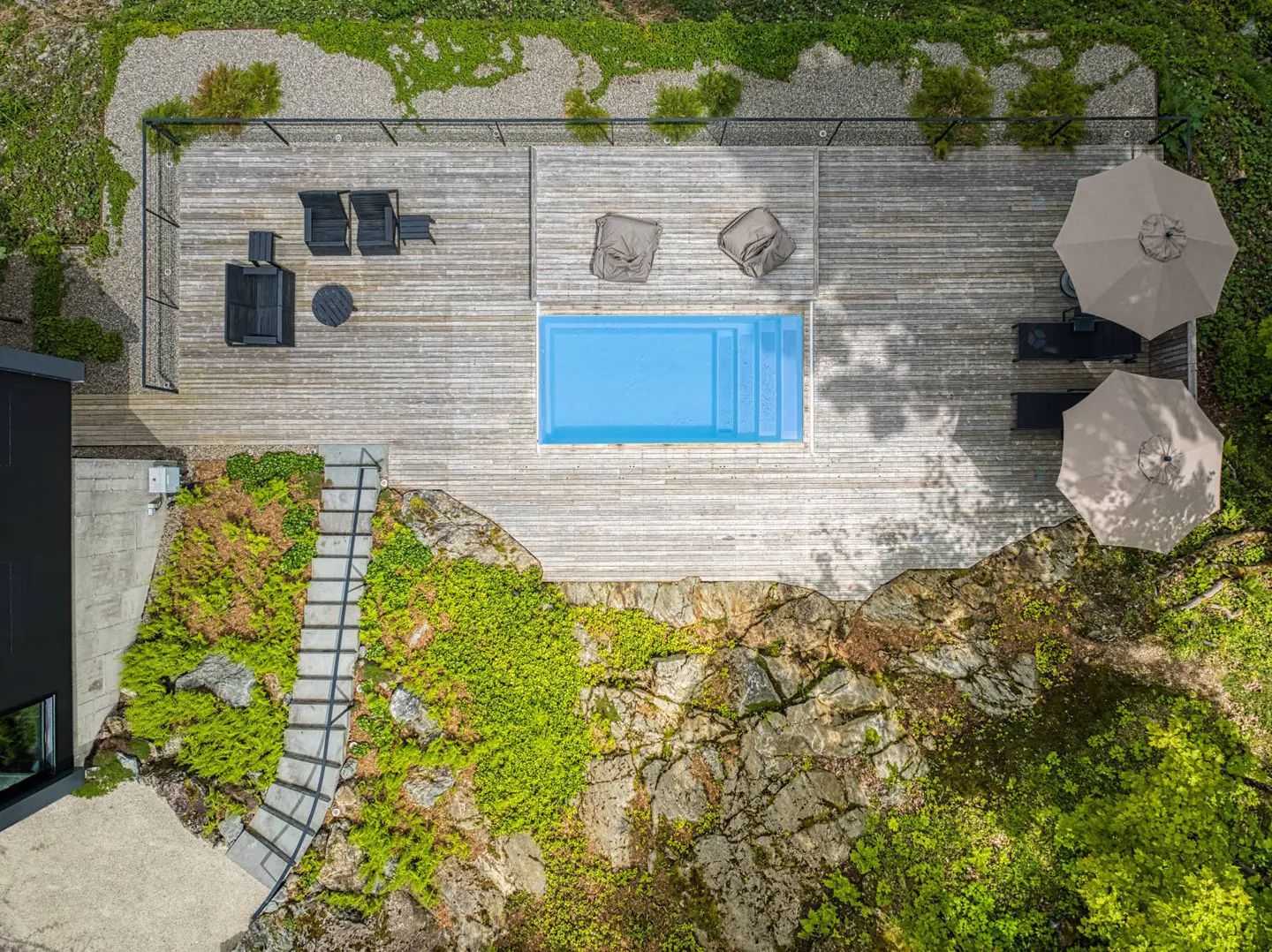 Aerial view of a wooden deck with a blue pool, lounge chairs, and umbrellas, surrounded by greenery and rock formations.