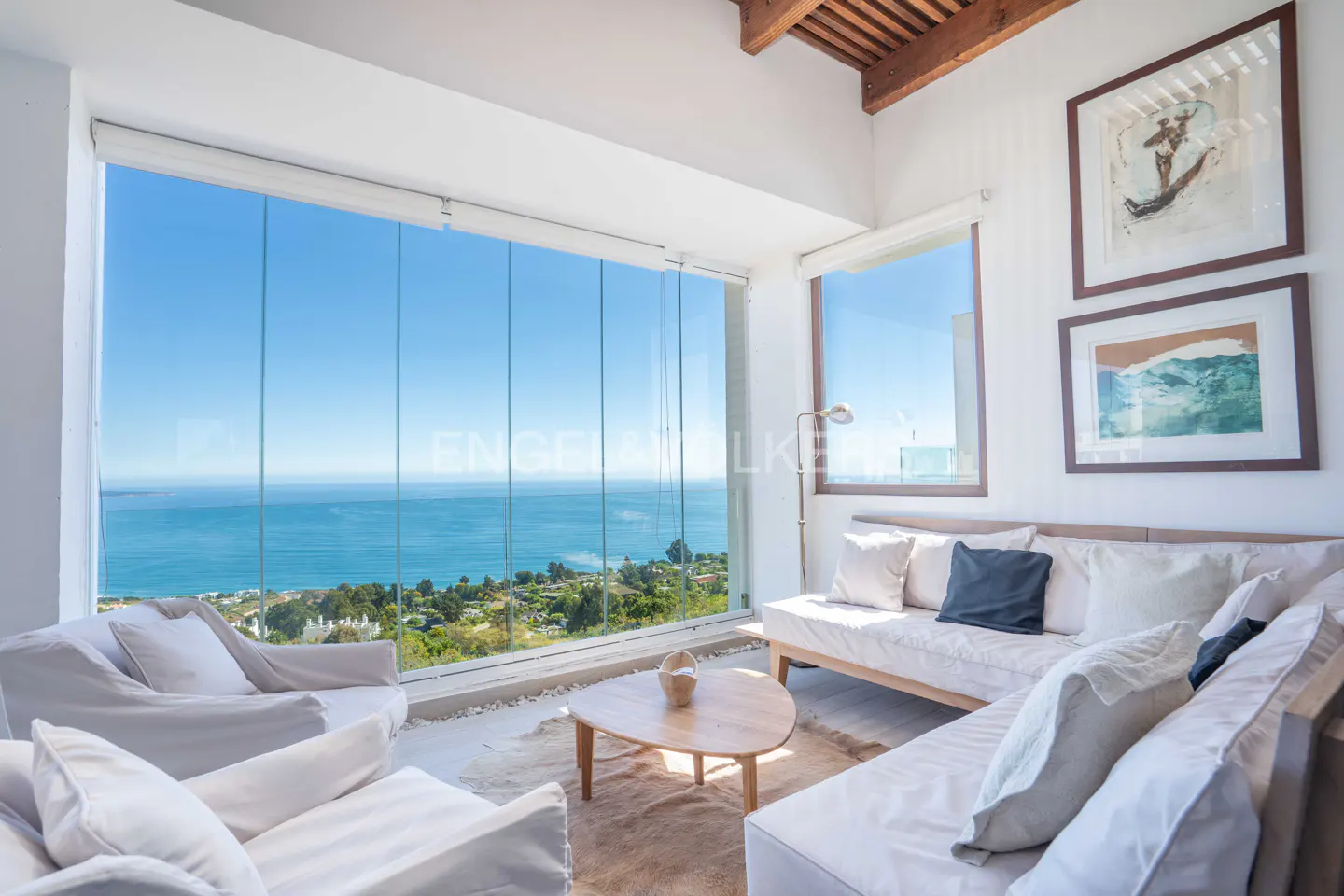 Bright living room with ocean view. White sofas and chairs face a large window overlooking the blue sea. Artwork hangs on the white walls.