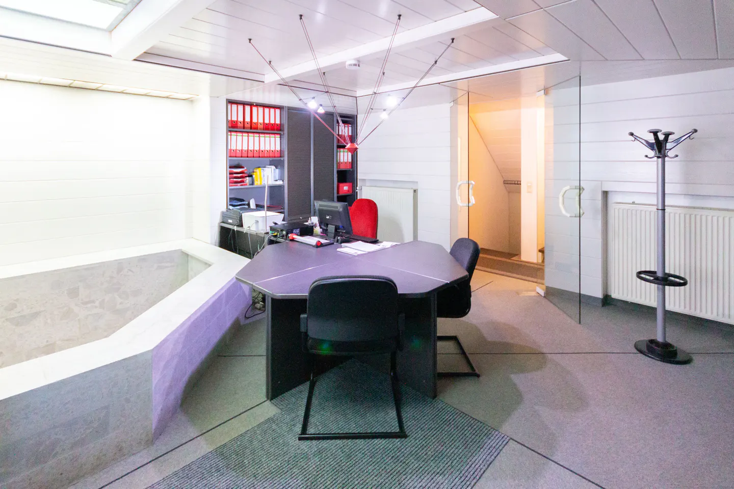 Office interior with a gray desk, black chairs, and a coat rack near a glass door. Red binders are on shelves in the background.
