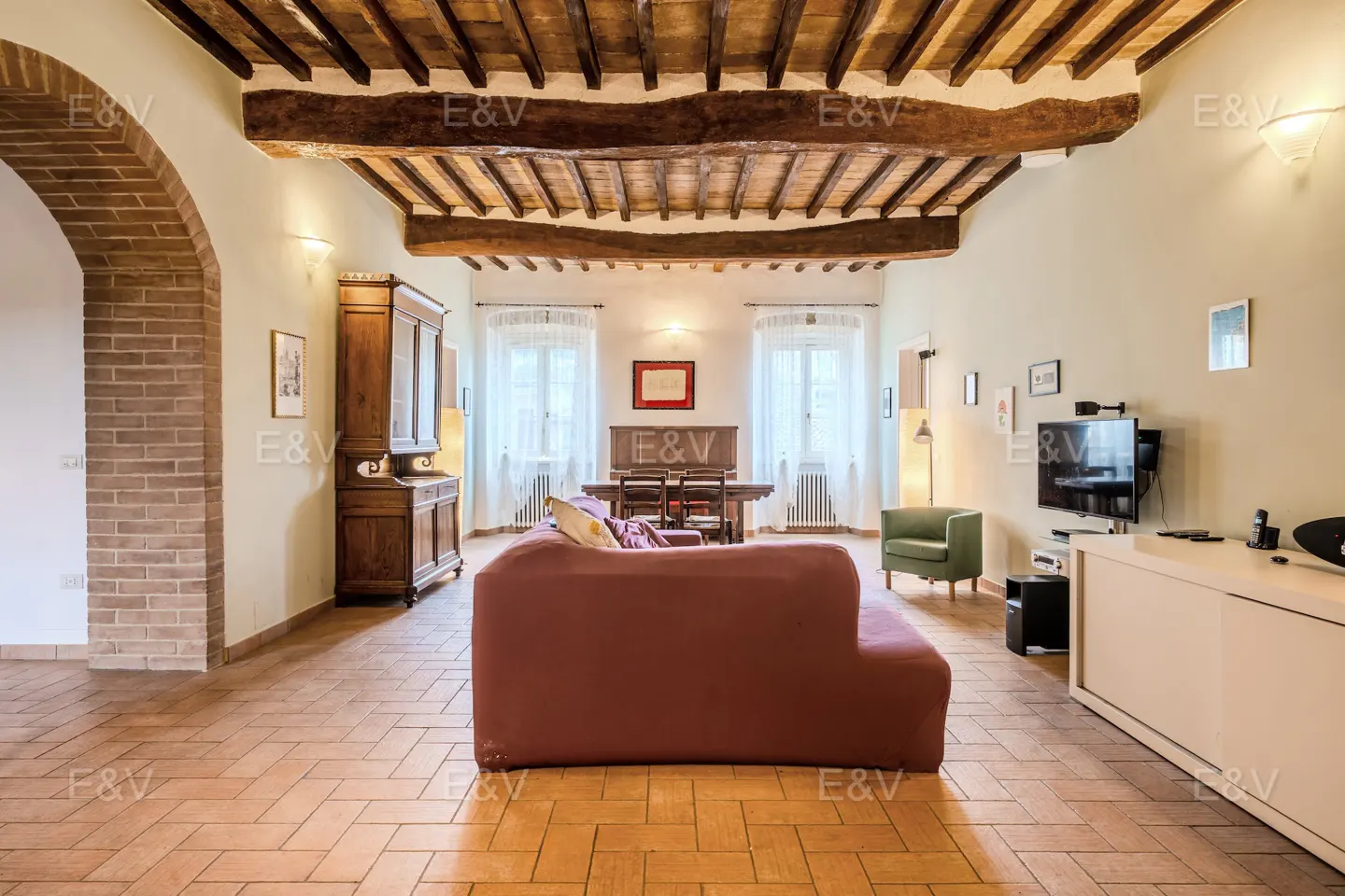 Living room with brick floors, exposed wood beam ceiling, red sofa, dining table, and TV.