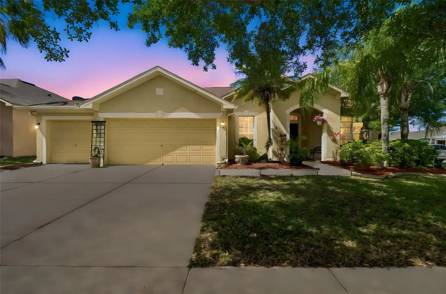 A tan single-story house with a two-car garage, green lawn, and palm trees at dusk.