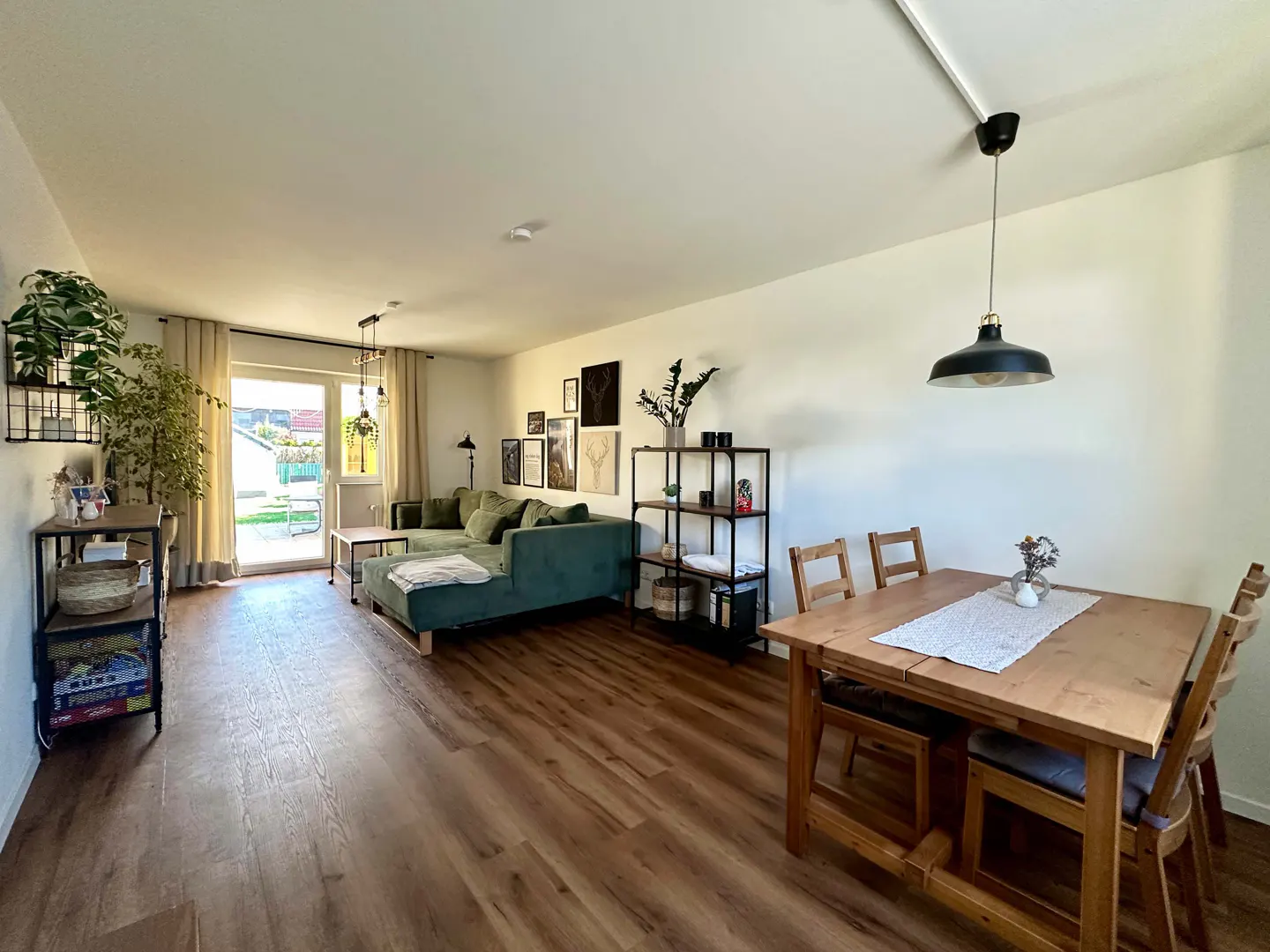 Bright, open-concept living and dining area with wood floors. A green sectional sofa faces a wood dining table with chairs. Shelves display plants and decor.