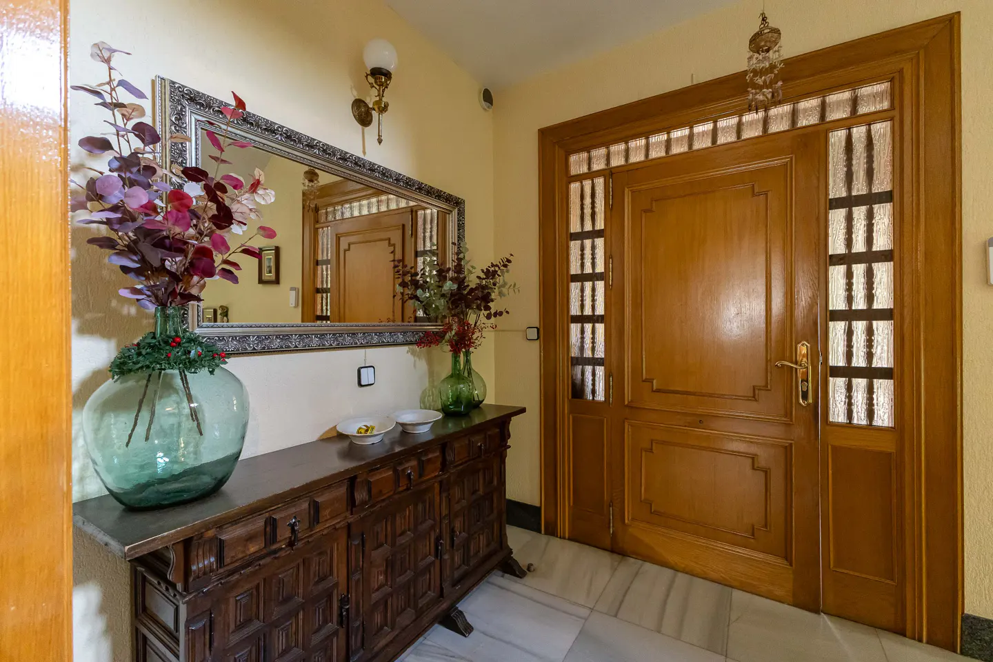 Entryway with a wooden door, marble floor, and a dark wood cabinet with a large mirror above it. A green vase with red flowers sits on the cabinet.