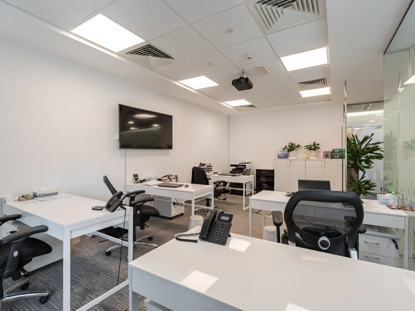 Modern office with white desks, black ergonomic chairs, a wall-mounted TV, and bright recessed lighting.