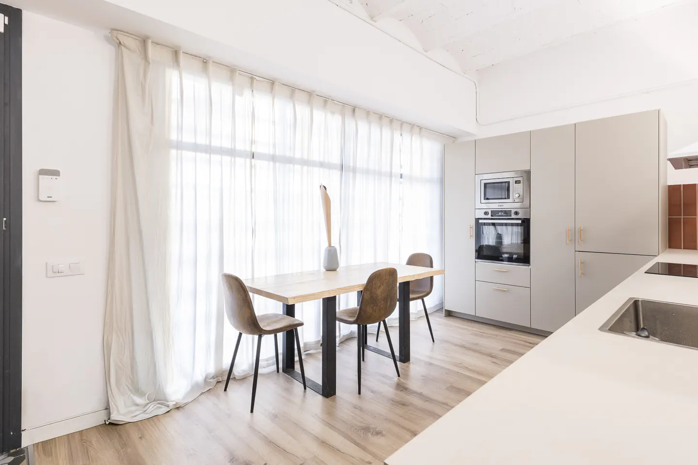 Bright kitchen with wood floors, a table with three chairs, and light gray cabinets. A large window with sheer white curtains is behind the table.