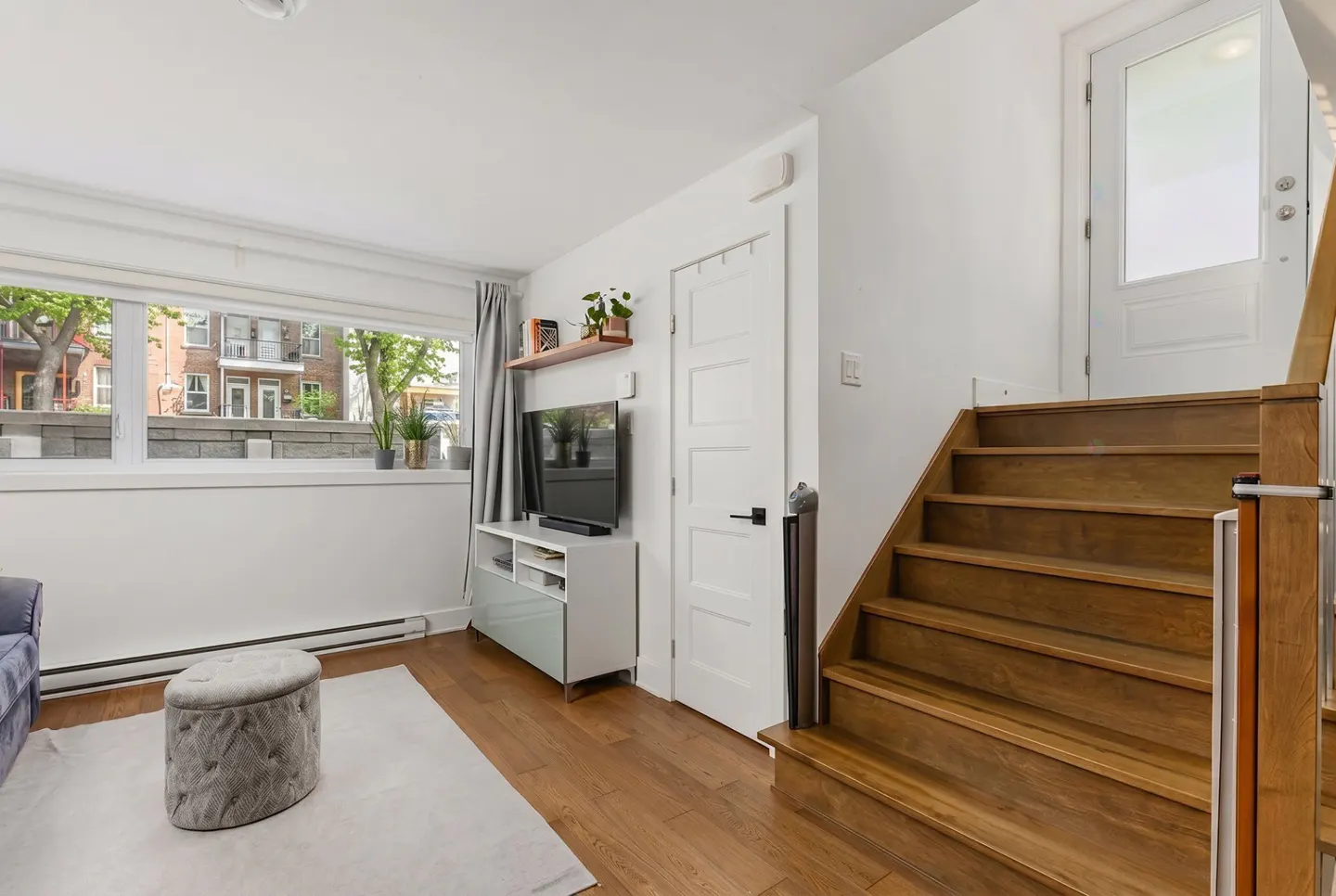 Bright living room with wood floors, white walls, and stairs. A window overlooks a city street. A TV sits on a white console.