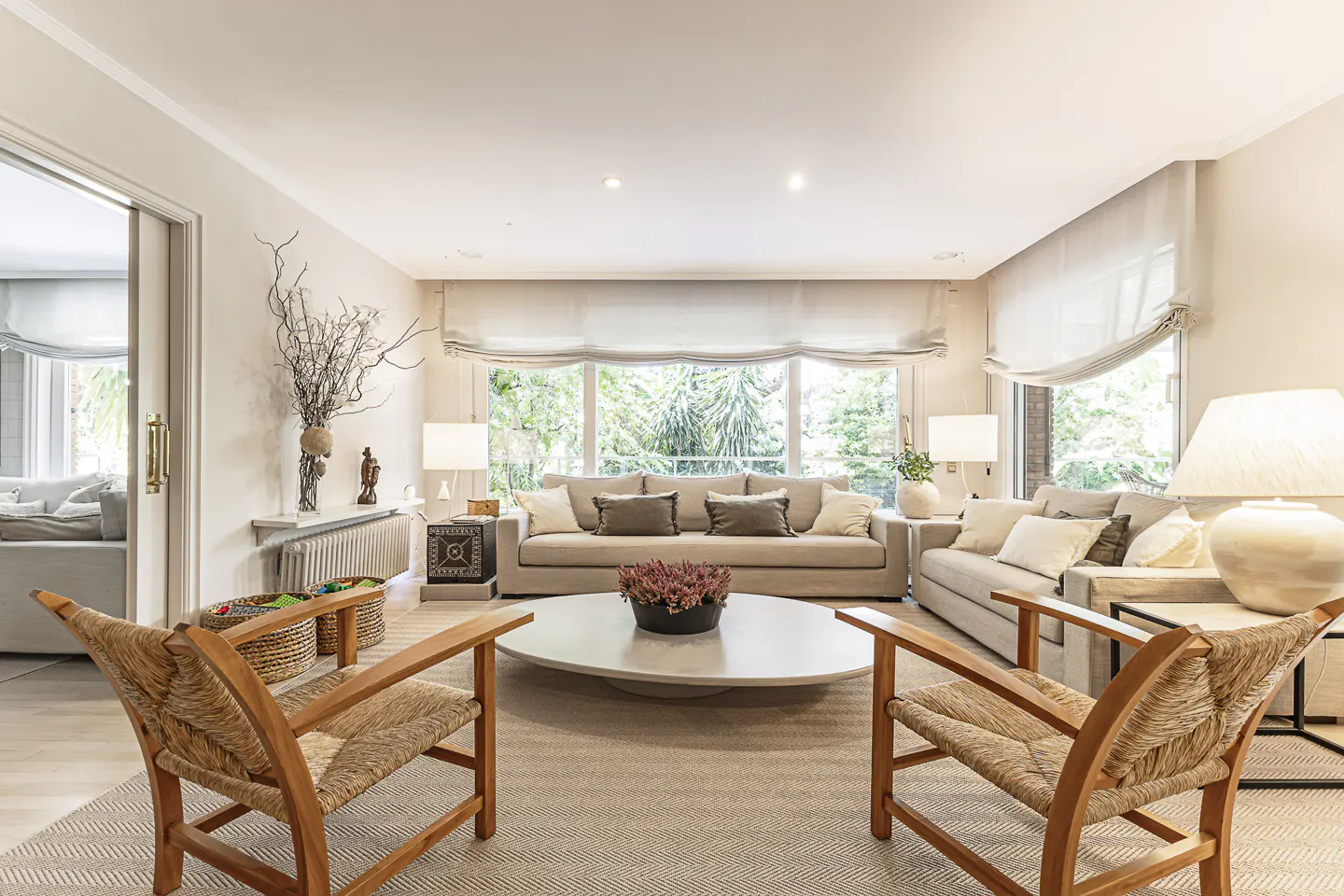 Living room with two sofas, a round table with flowers, and two wooden chairs on a herringbone rug. Large window with beige blinds.