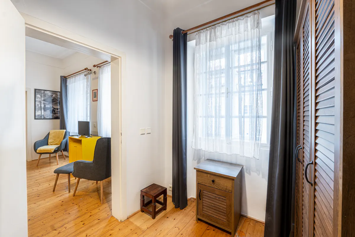 A room with wooden floors, a window with white curtains, and a dark wardrobe. A doorway leads to another room with a yellow desk and chairs.