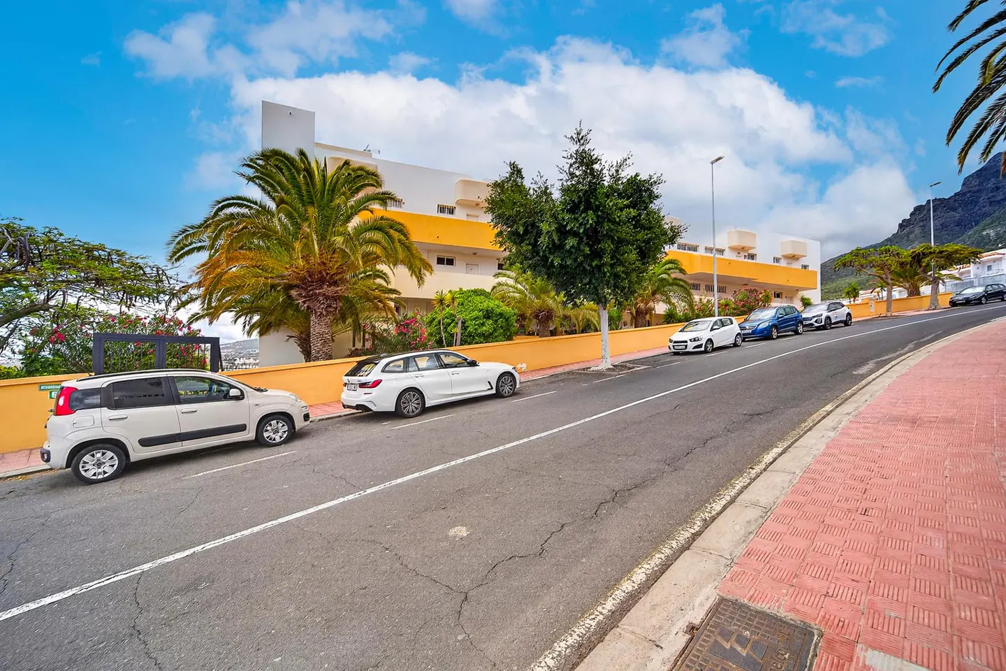Street view of a white building with yellow accents, palm trees, and parked cars under a blue, cloudy sky.