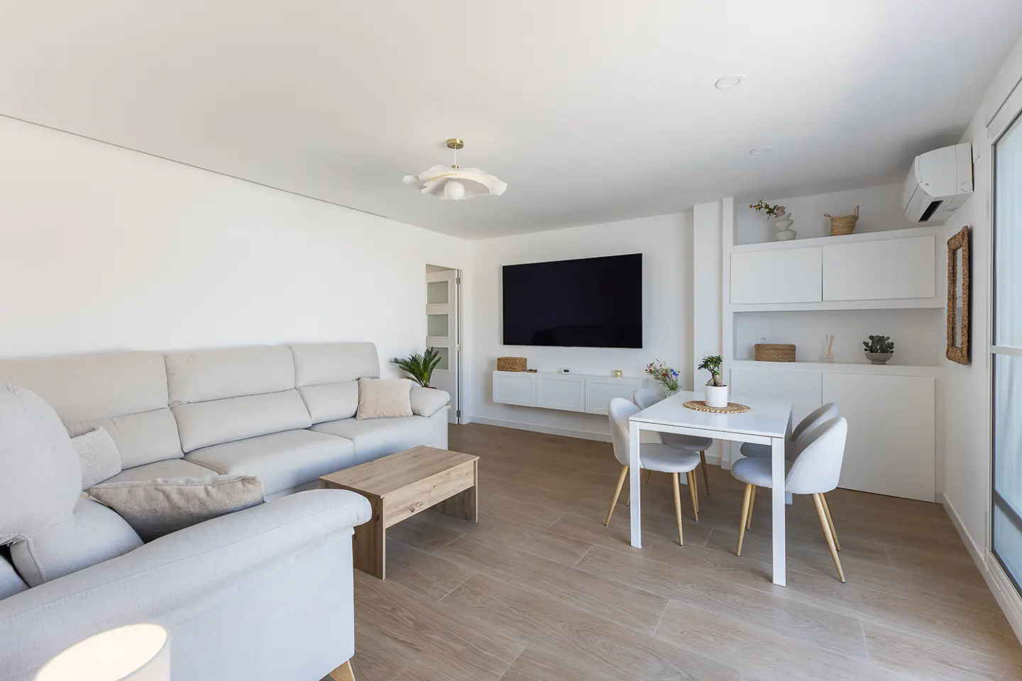 Bright living room with a beige sofa, wood coffee table, and a white dining table with four chairs. A large TV hangs on the white wall.