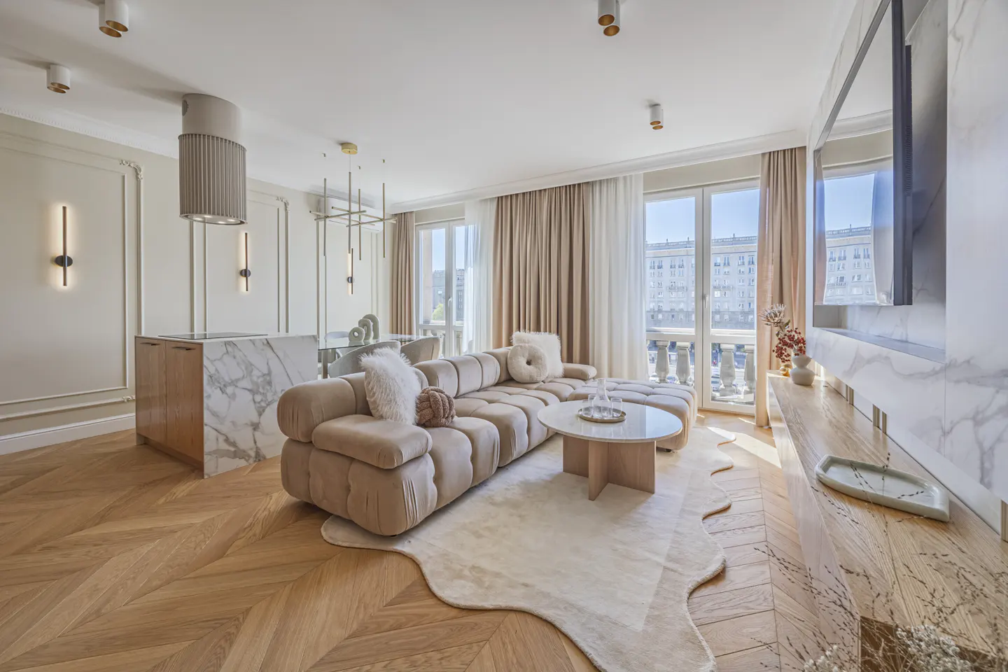 Bright living room with herringbone wood floors, a beige sectional sofa, and marble accents. Large windows offer city views.