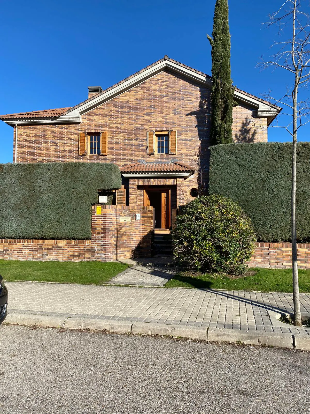 Two-story brick house with brown shutters and a red tile roof, surrounded by green hedges and trees.