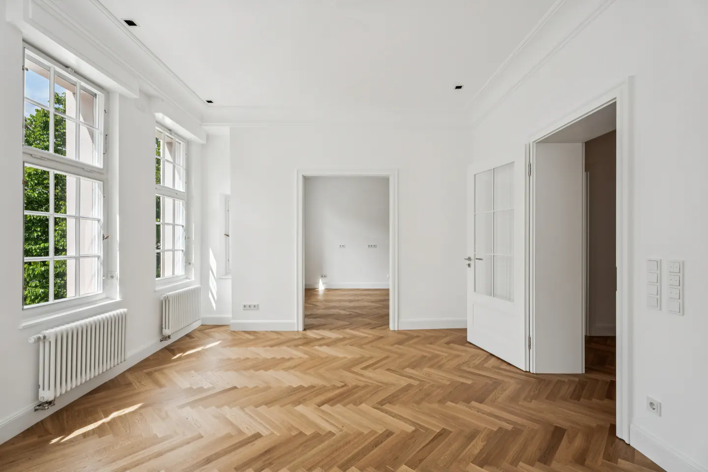 Bright, empty room with herringbone wood floors, white walls, and large windows. Doorways lead to other rooms. Radiator under windows.