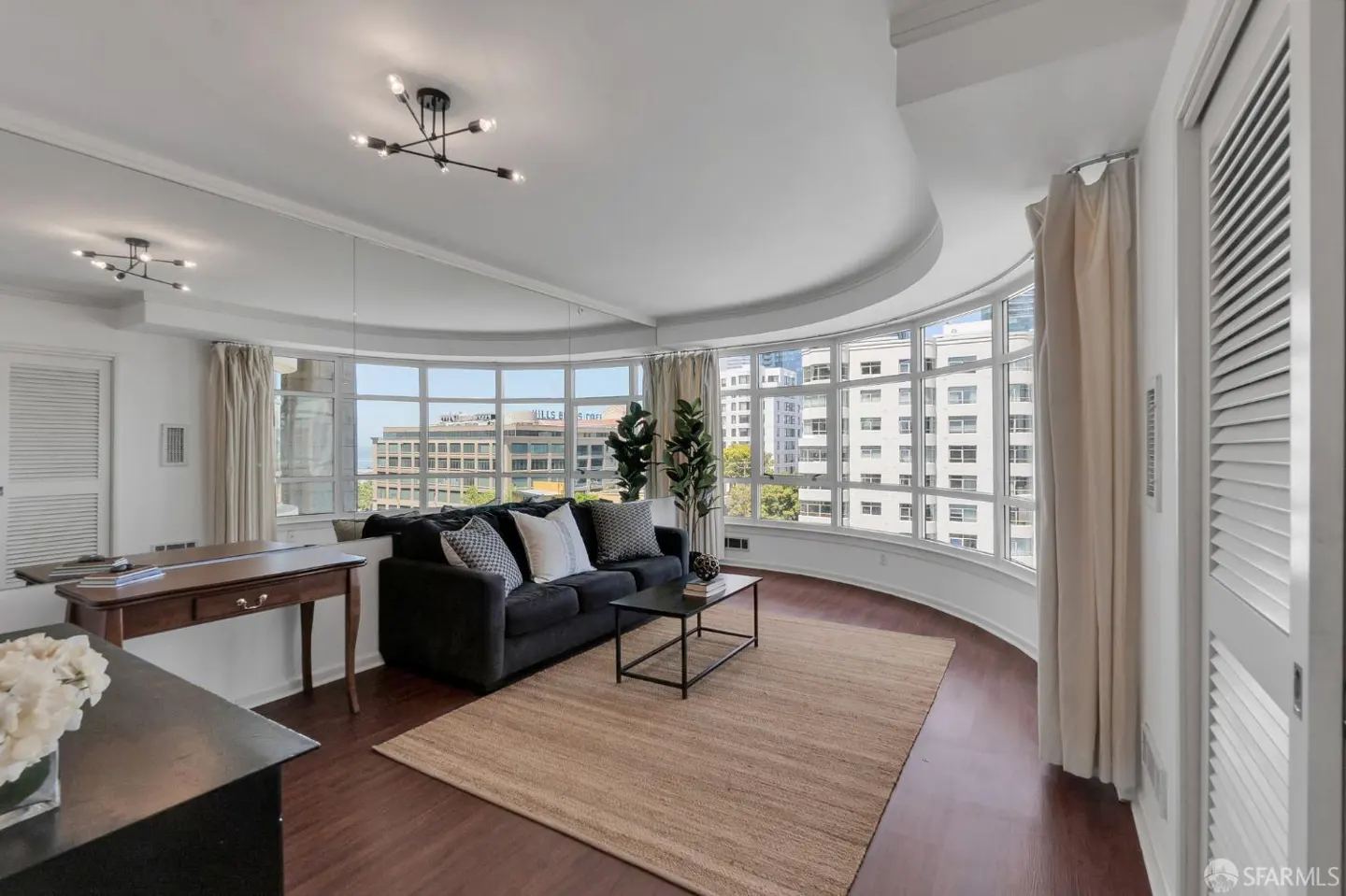 Bright living room with curved window wall, dark sofa, and tan rug. City buildings are visible through the windows.