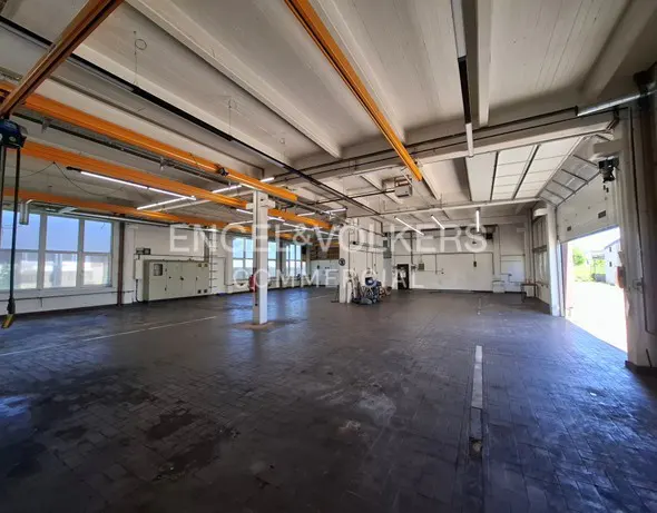 Spacious industrial warehouse with a dark tiled floor, white walls, large windows, and yellow overhead crane beams.