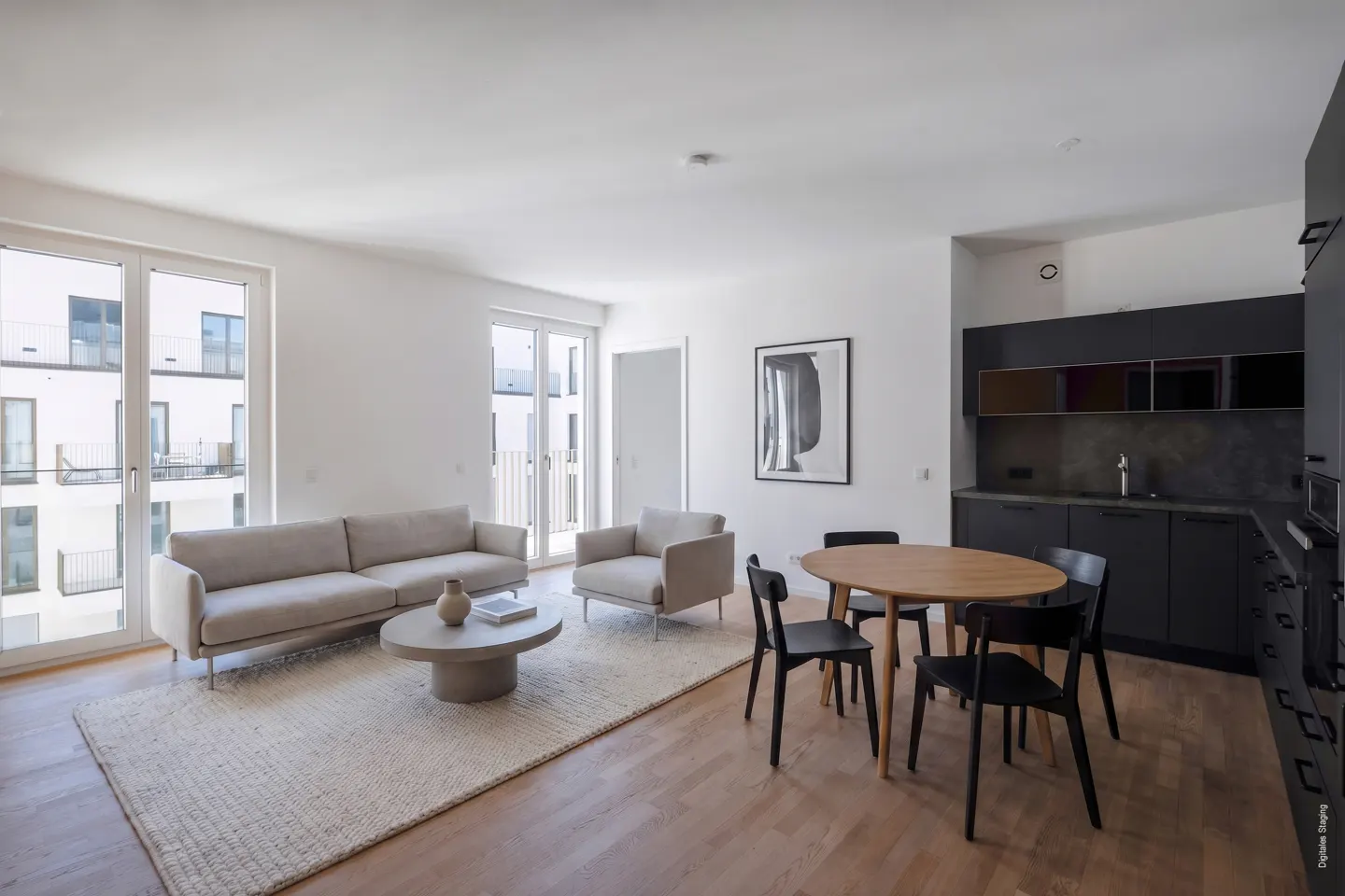 Bright, modern apartment with a beige sofa, round coffee table, and a wooden dining table with black chairs. Black kitchen cabinets in the background.