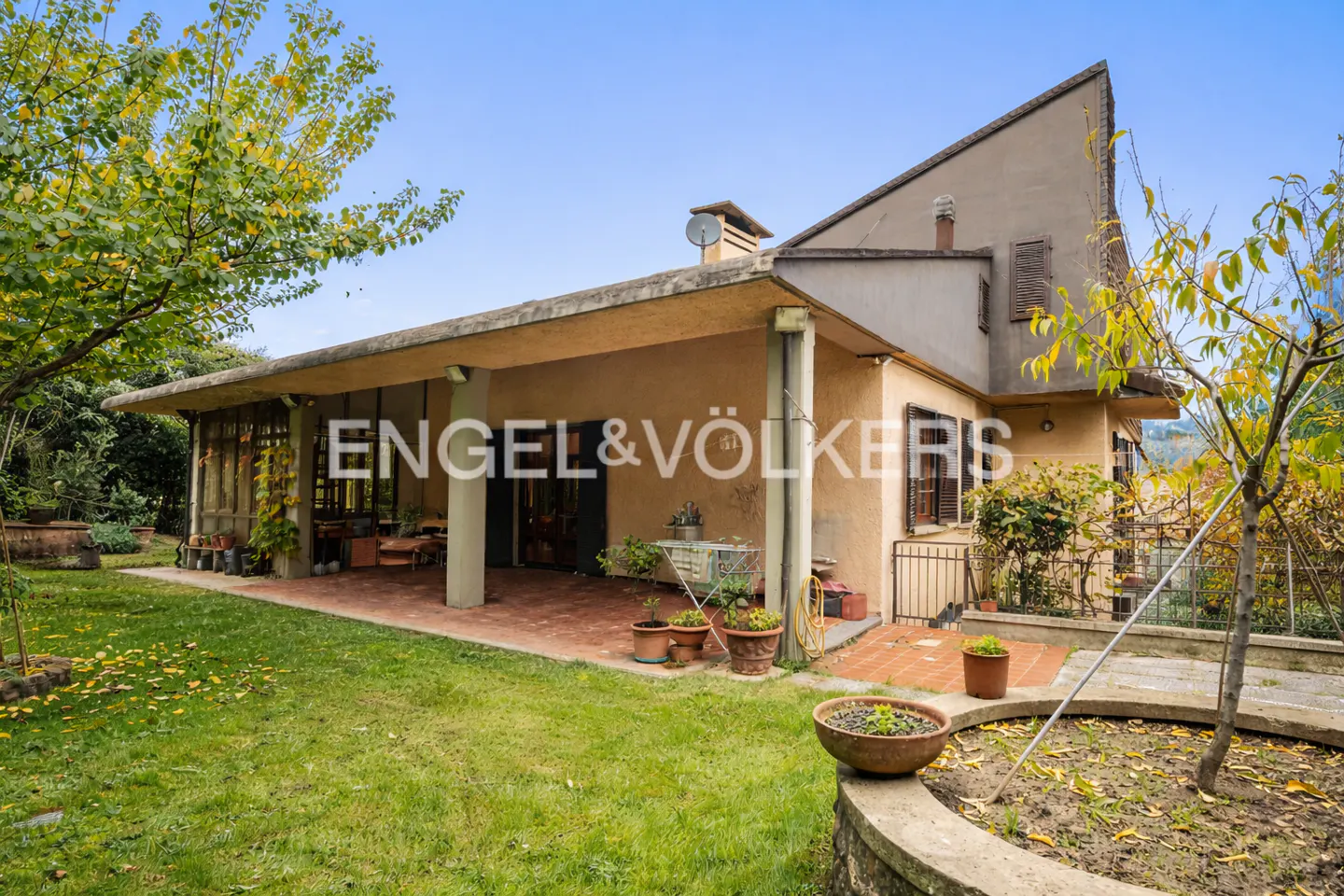 Exterior view of a beige two-story house with a covered patio, green lawn, and trees under a blue sky. Engel & Völkers logo is visible.