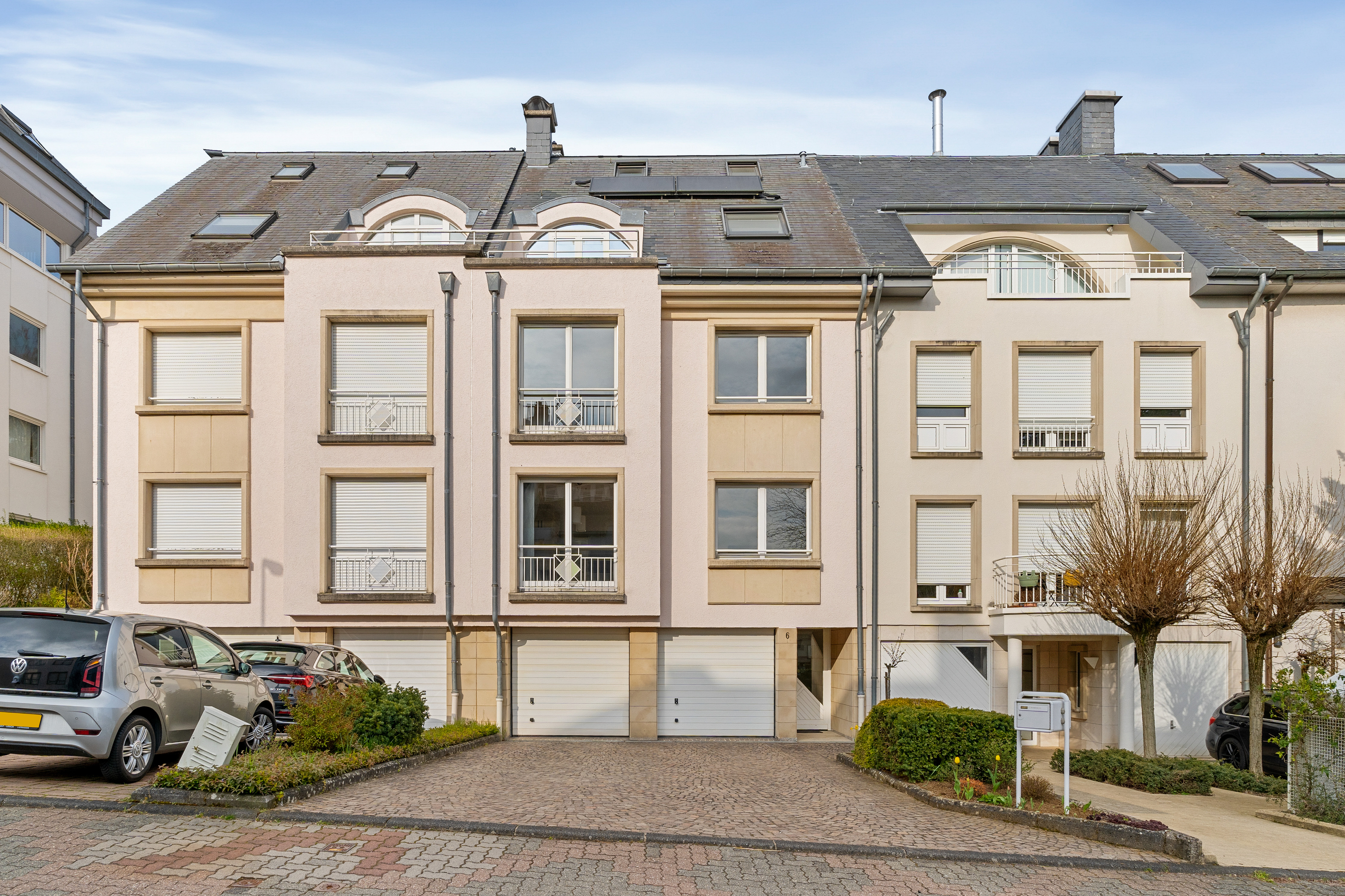 Row of three-story townhouses with garages. The buildings are light beige with gray roofs and dormer windows. Cars are parked in front.