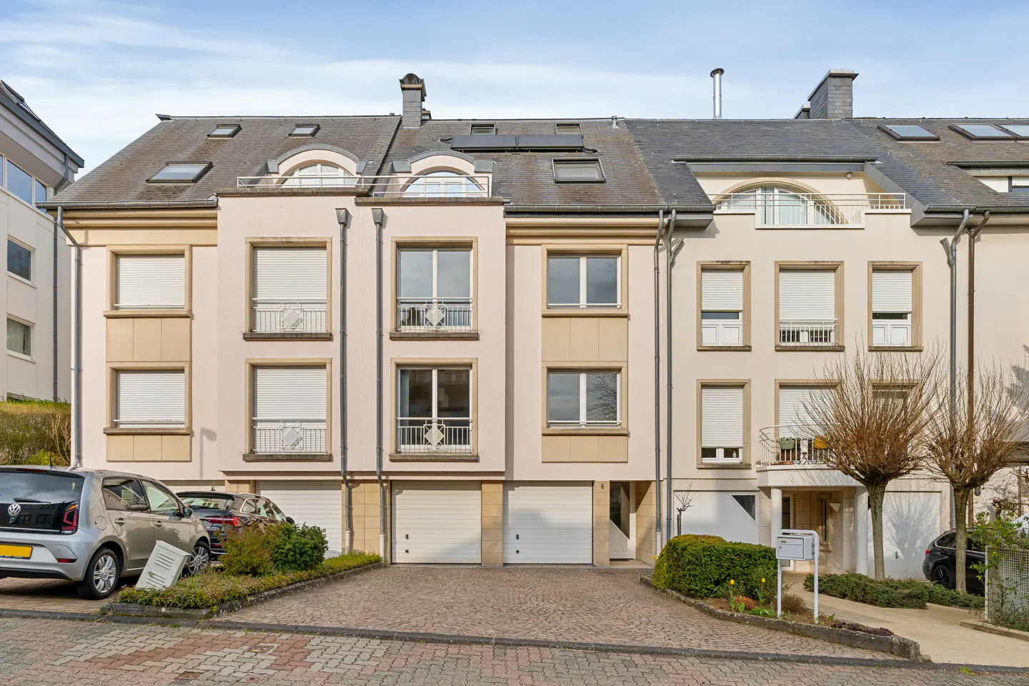 Row of three-story townhouses with garages. The buildings are light beige with gray roofs and dormer windows. Cars are parked in front.