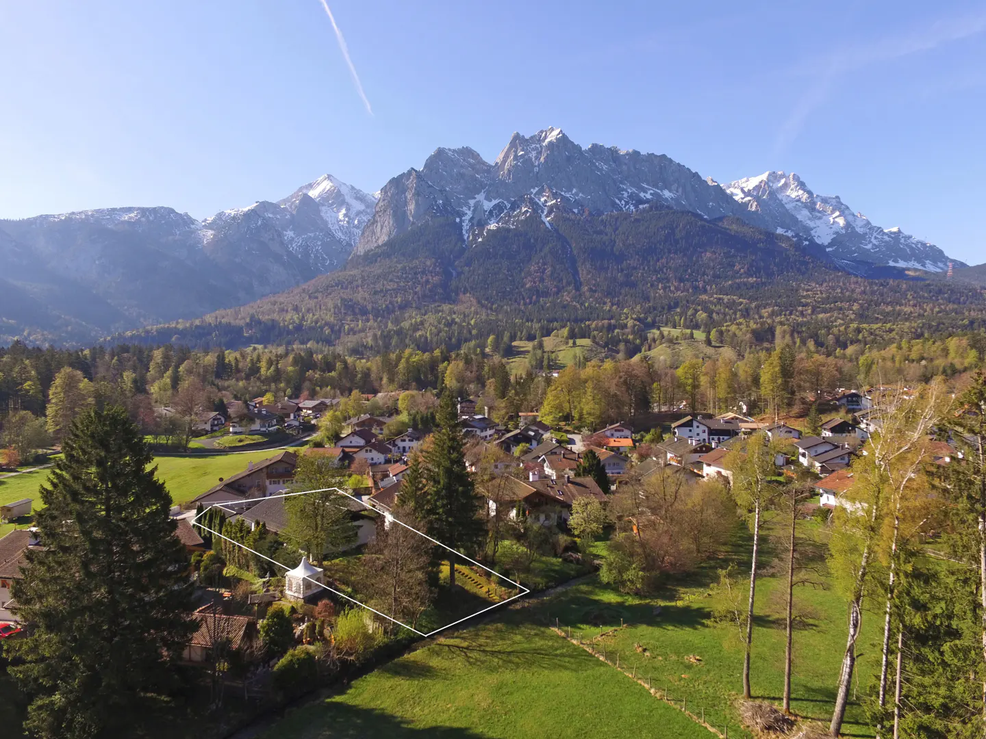 Aerial view of a property outlined in white, nestled in a village with snow-capped mountains in the background.