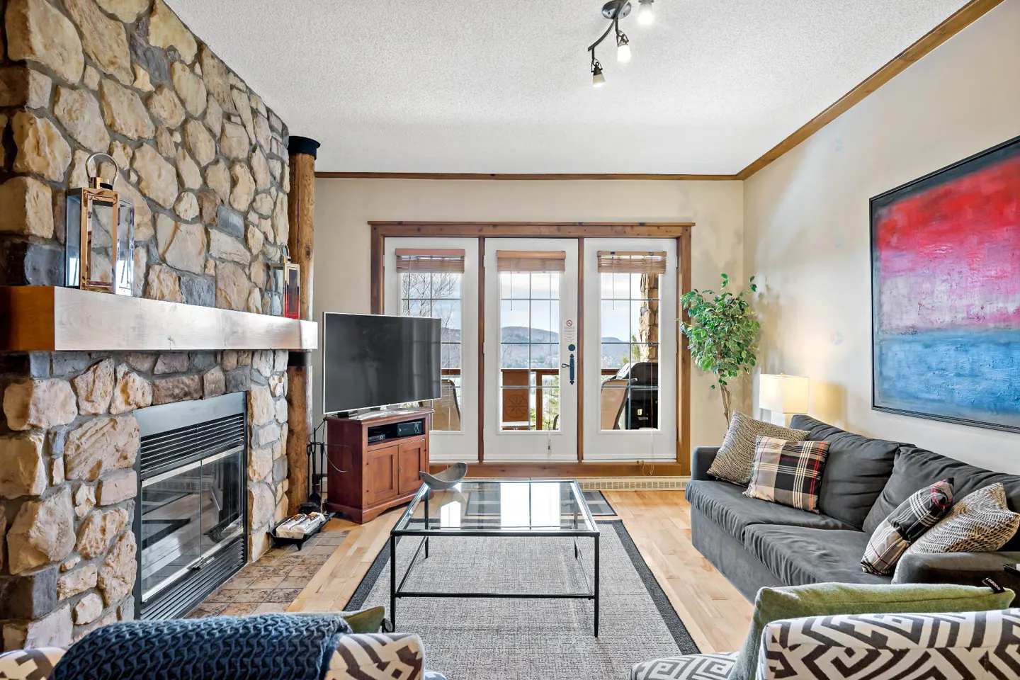 Living room with stone fireplace, gray sofa, glass coffee table, and doors to a balcony with mountain views.
