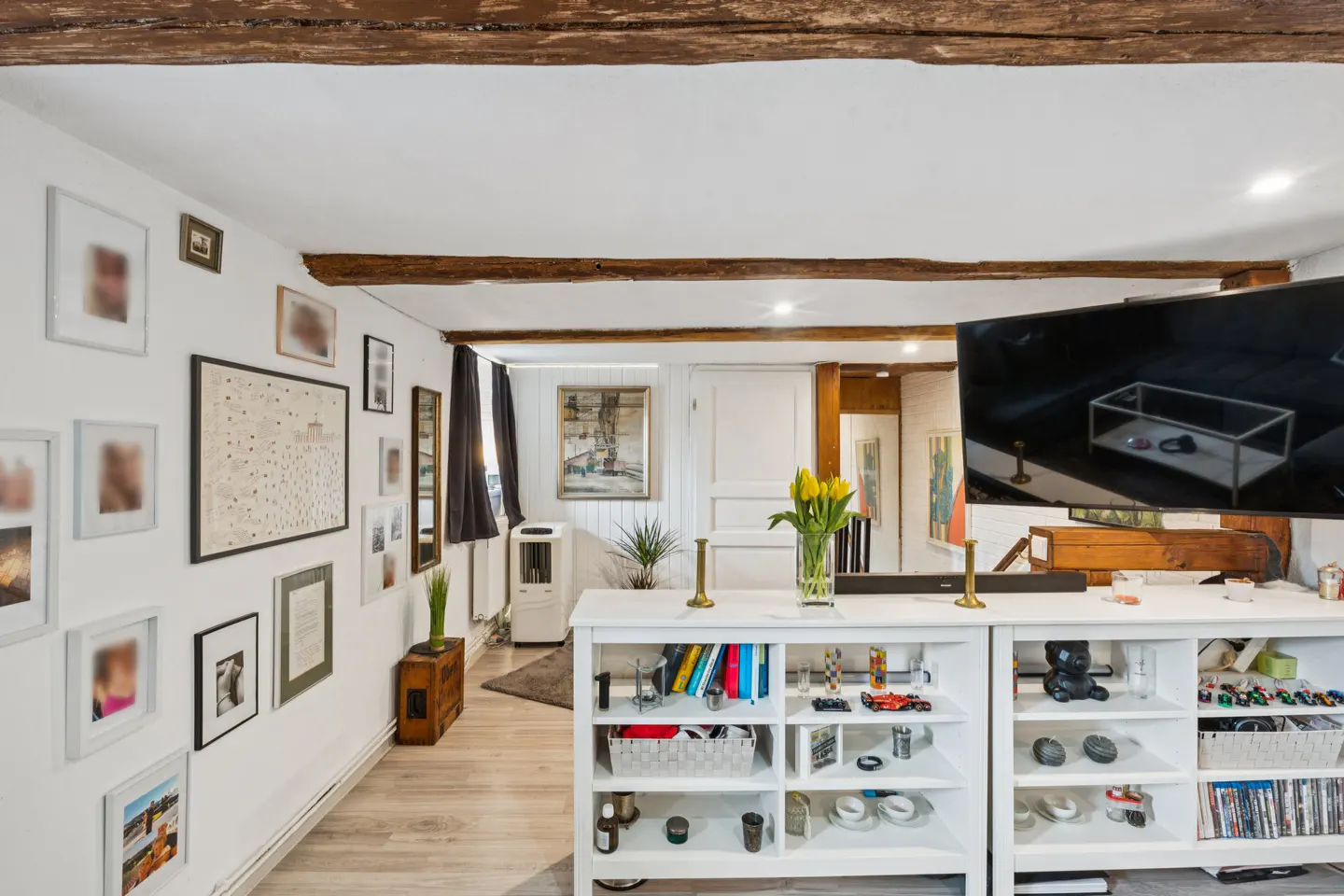 Living room with white walls, wood floors, and exposed beams. A white shelving unit holds books and decor. A TV is mounted above.