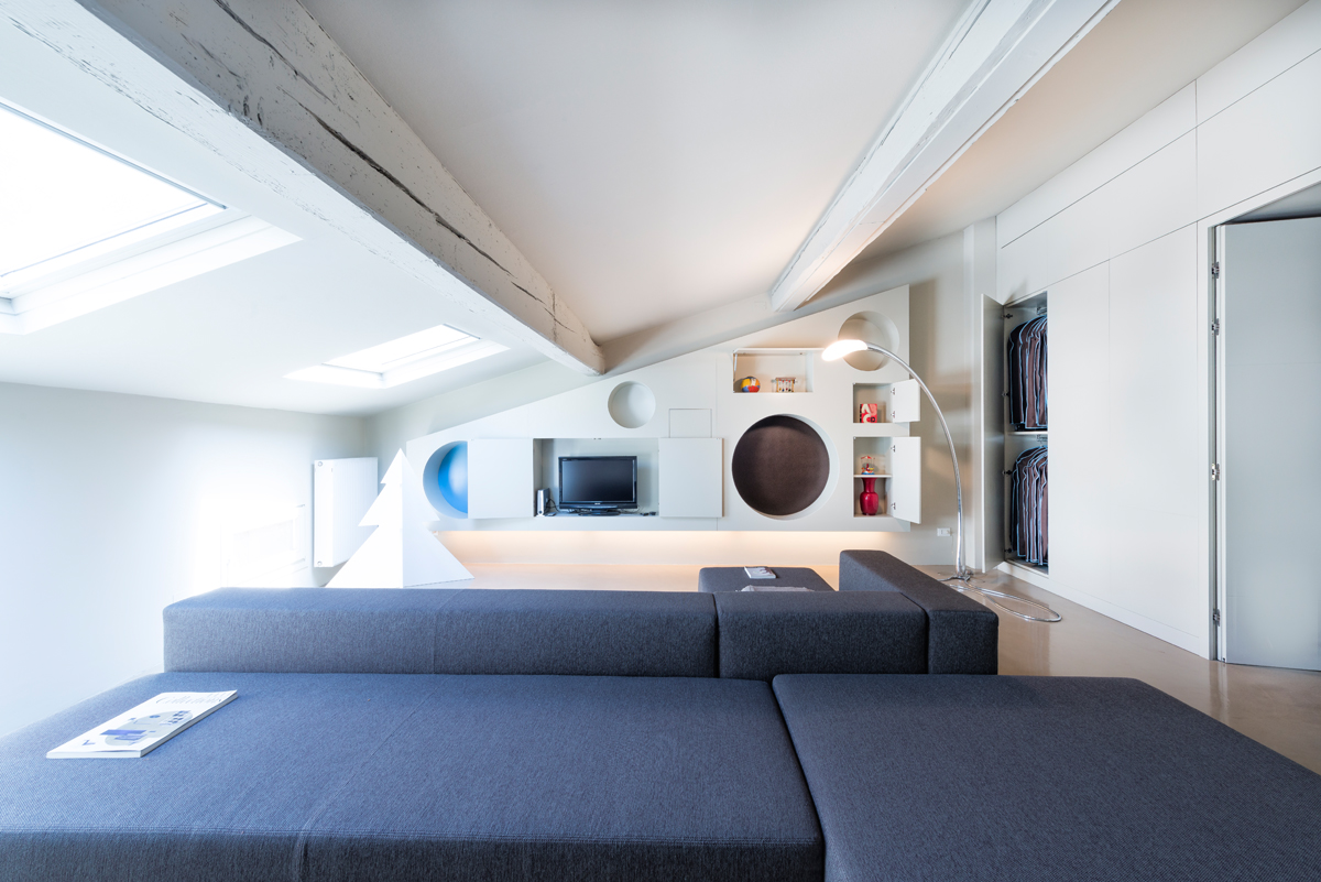 Attic living room with a gray sectional sofa, white walls, and built-in shelving with circular cutouts. Skylights and exposed beams add architectural detail.