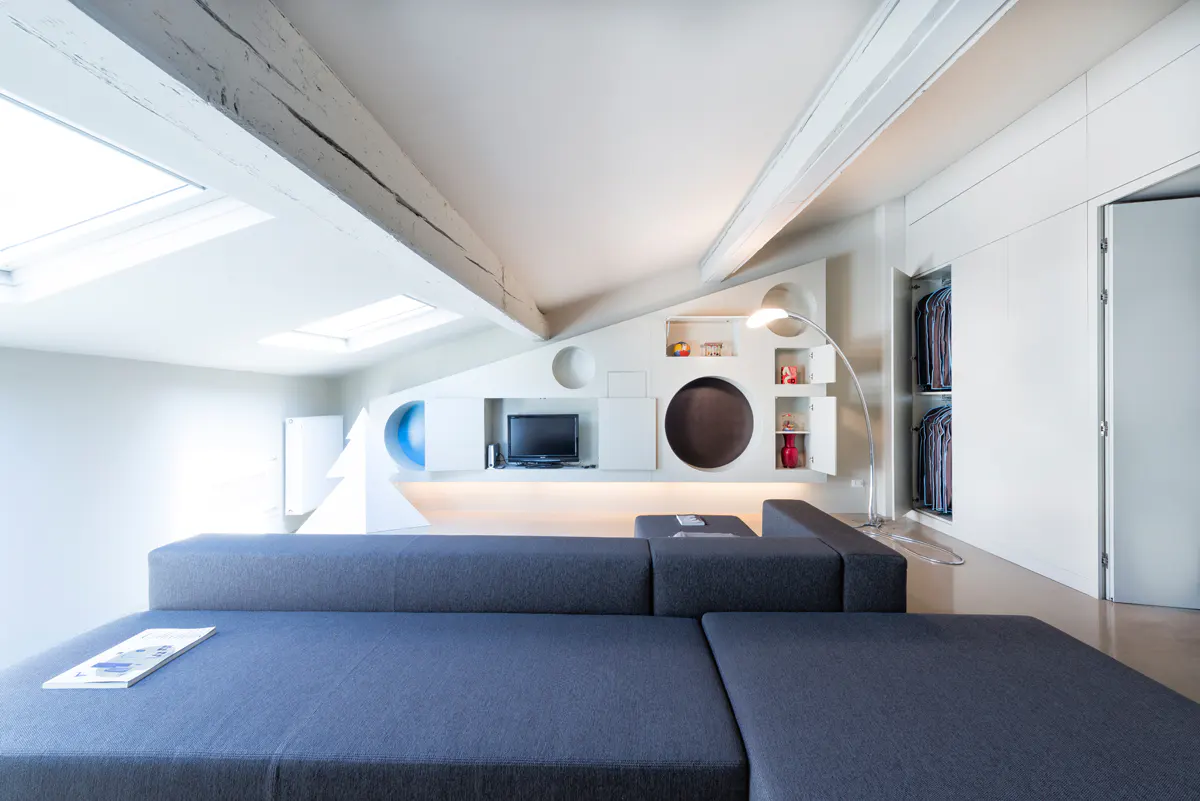 Attic living room with a gray sectional sofa, white walls, and built-in shelving with circular cutouts. Skylights and exposed beams add architectural detail.