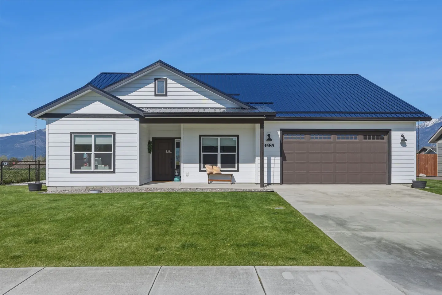 A white, one-story house with a blue metal roof, brown garage door, and green lawn on a sunny day.