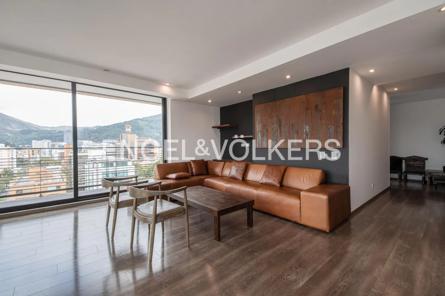 Modern living room with a tan leather sectional, wooden coffee table, and two chairs, featuring a city view through windows.
