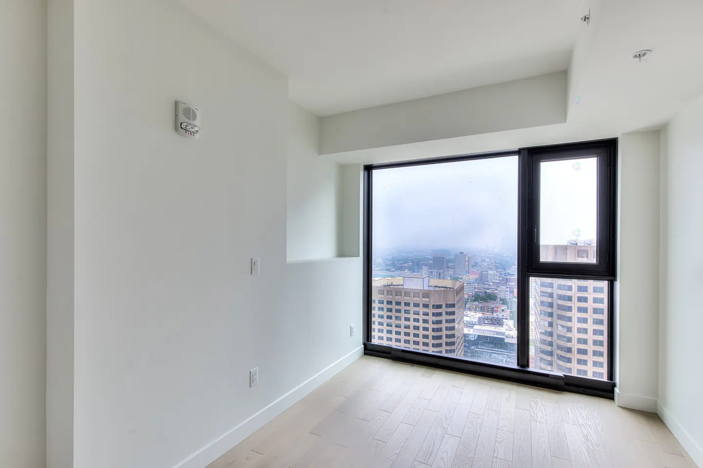Bright, empty room with white walls and light wood floors. A large black-framed window shows a city skyline on a cloudy day.