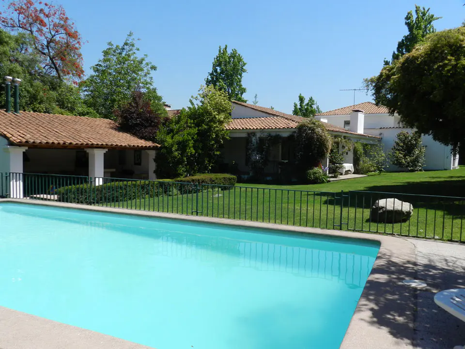 A turquoise pool is in front of a white house with a red tile roof, green lawn, and trees under a blue sky.