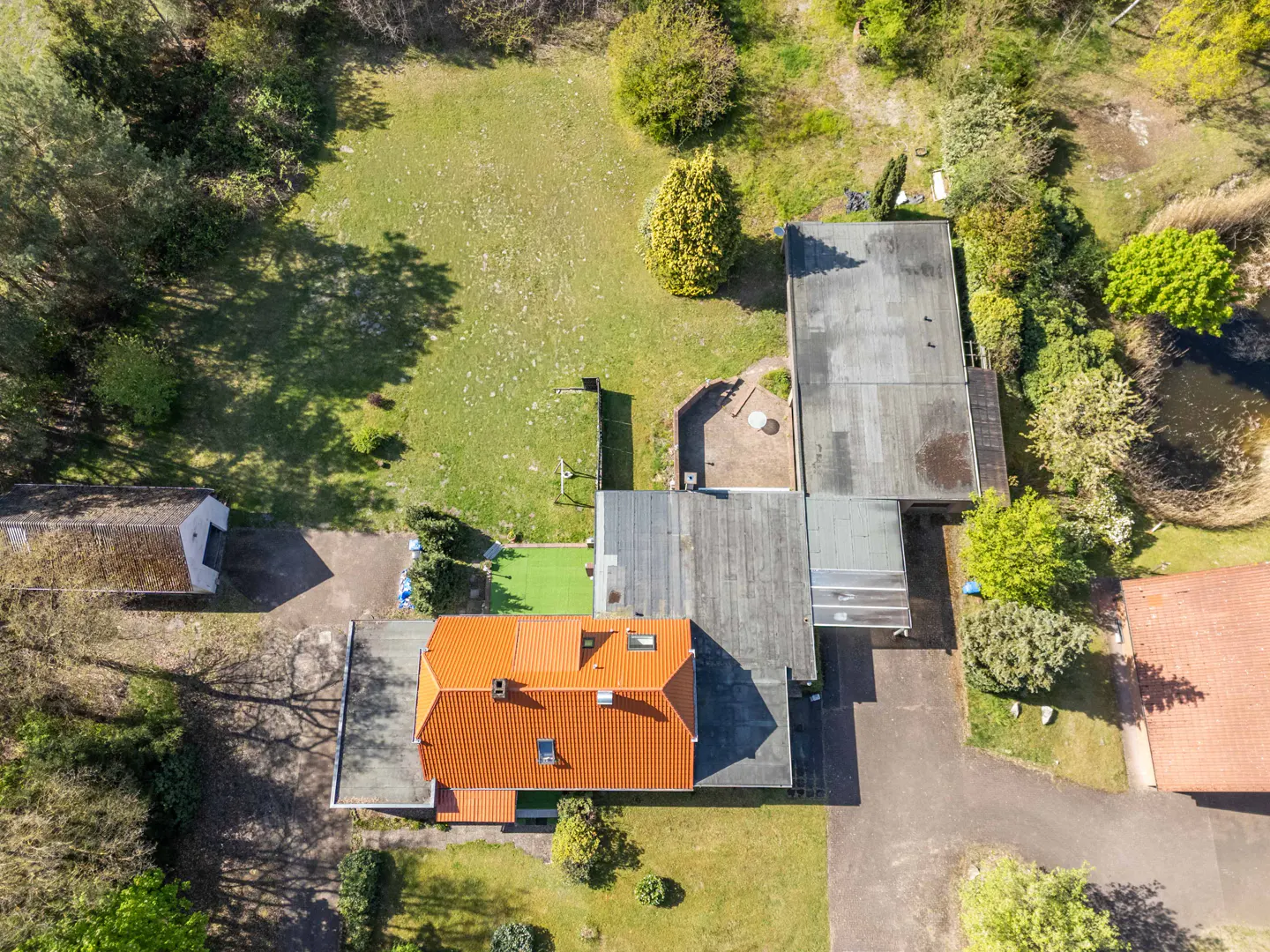 Aerial view of a house with an orange roof, surrounded by green grass, trees, and a pond. A gray driveway leads to the house.