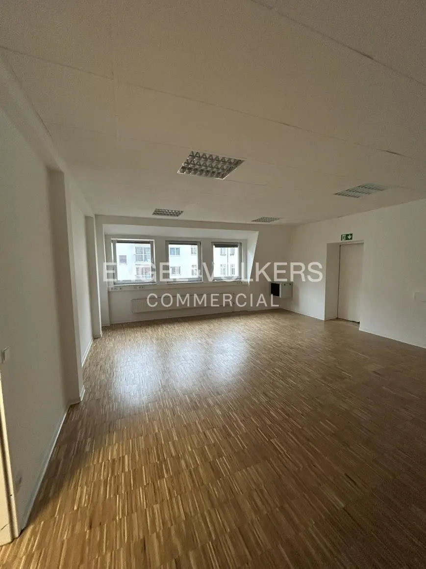 Bright, empty office space with parquet floors, white walls, and three windows. Overhead lighting and an exit door are visible.