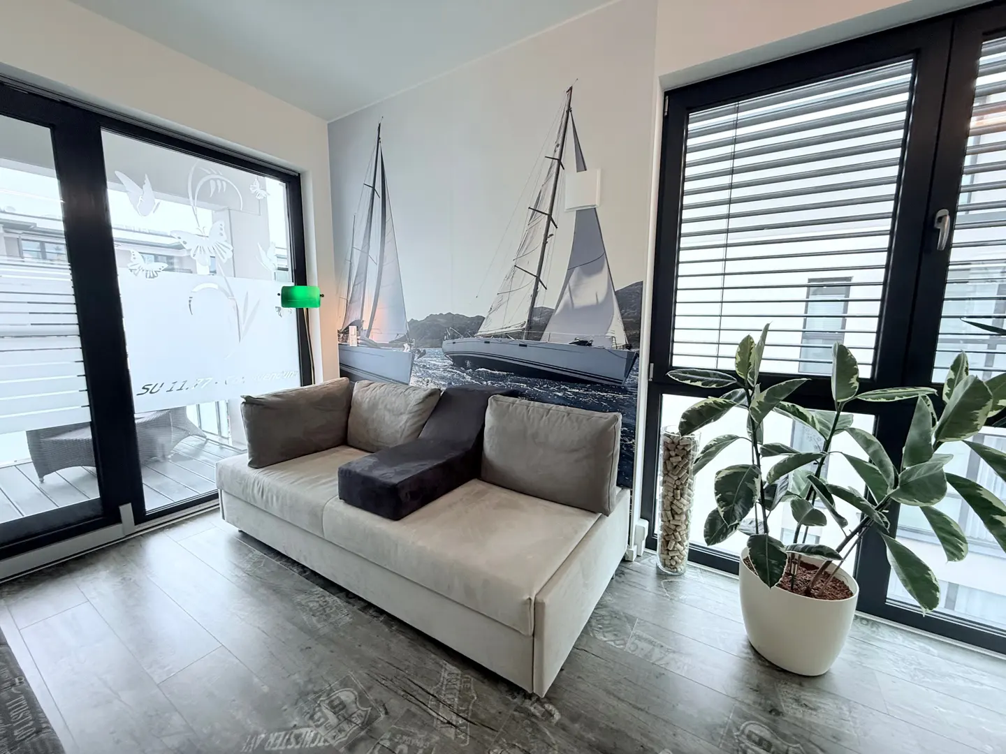Living room with a beige sofa, dark pillows, and a sailboat mural on the wall. A potted plant sits by the window.