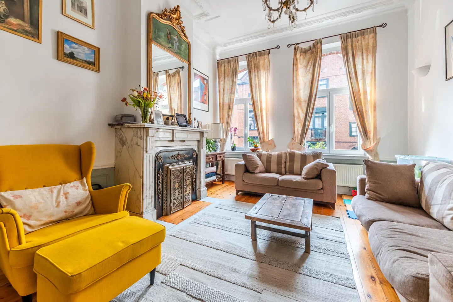 Bright living room with a yellow armchair, beige sofas, and a marble fireplace with a large ornate mirror above.