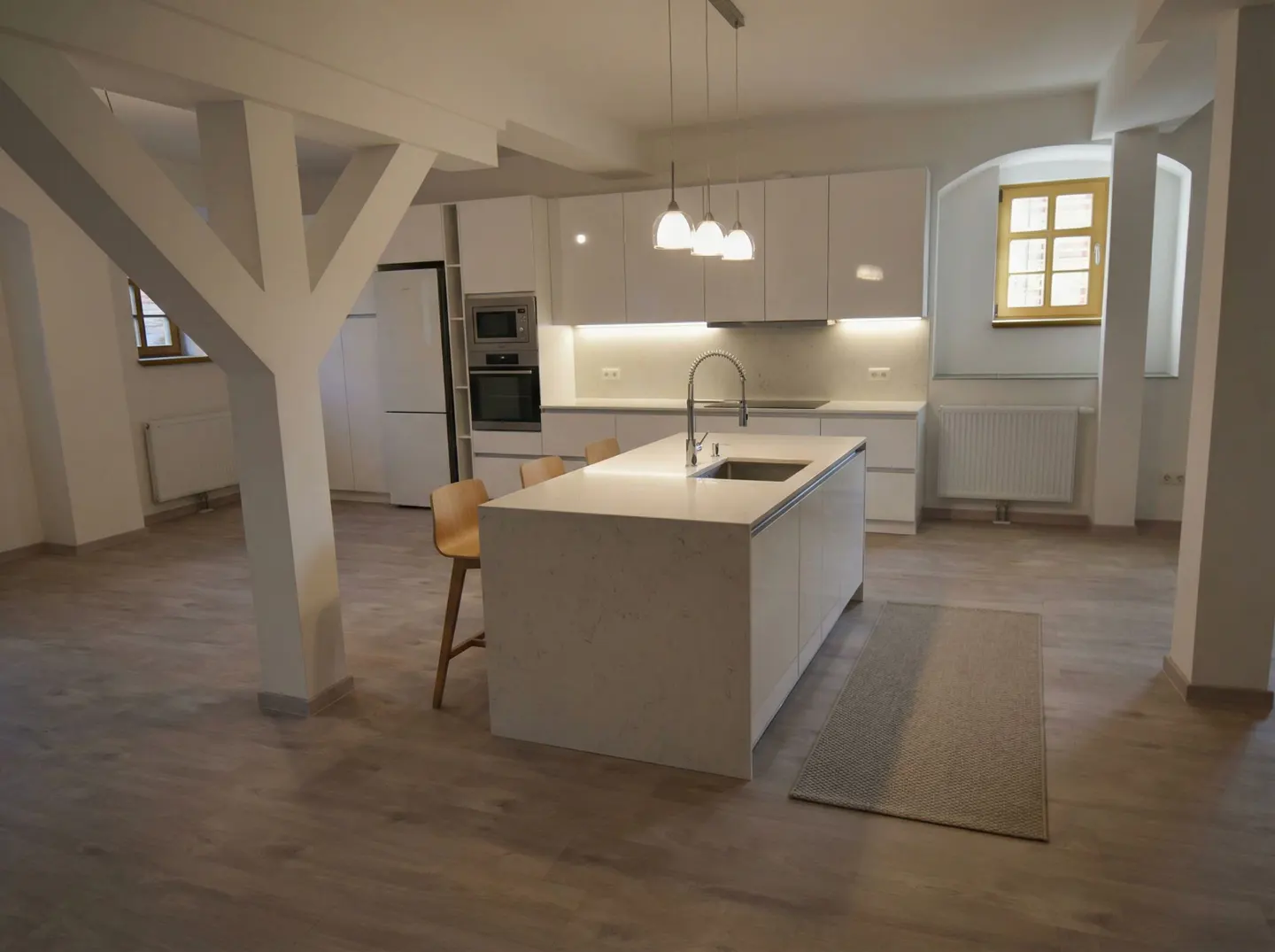 Bright, modern kitchen with white cabinets, island, and wood floors. Three pendant lights hang above the island. Large support beams are visible.