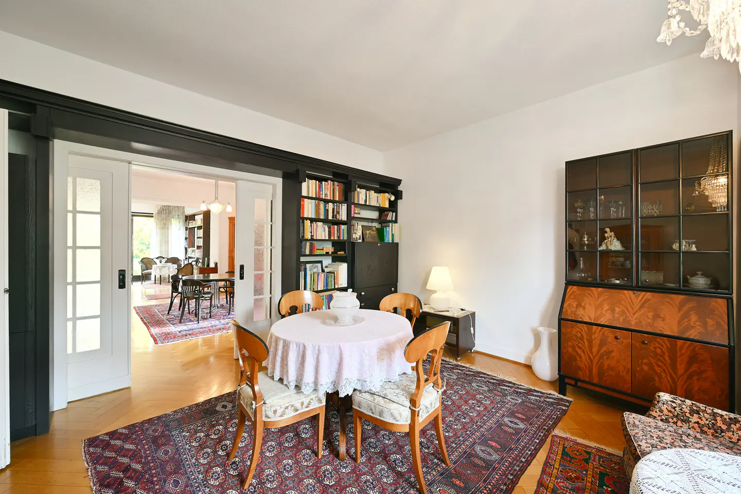 A bright room with a round table, chairs, a bookcase, and a glass cabinet. A rug covers the wooden floor. Sliding doors lead to another room.
