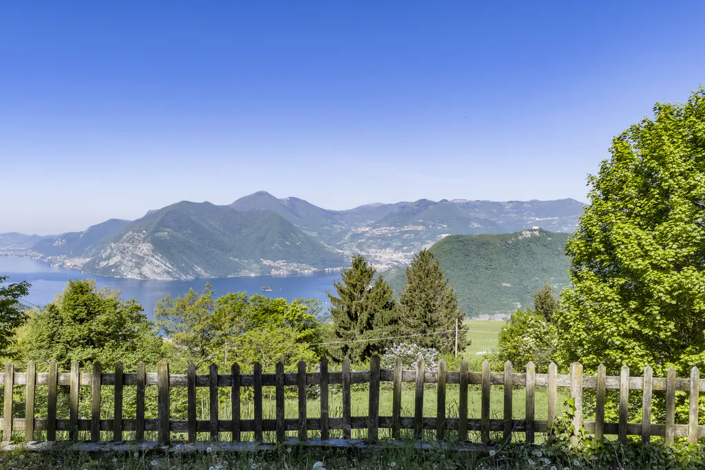 Scenic view of a lake and mountains under a blue sky, framed by a wooden fence and green trees.