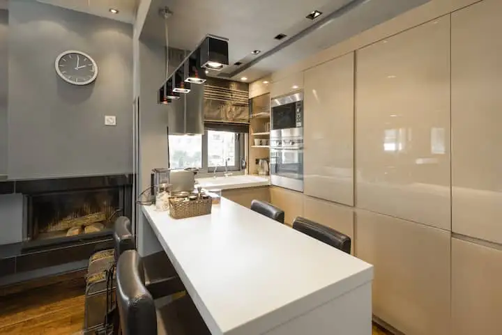 A modern kitchen with a white countertop island and black leather chairs. A fireplace is on the left, and beige cabinets and appliances are on the right.