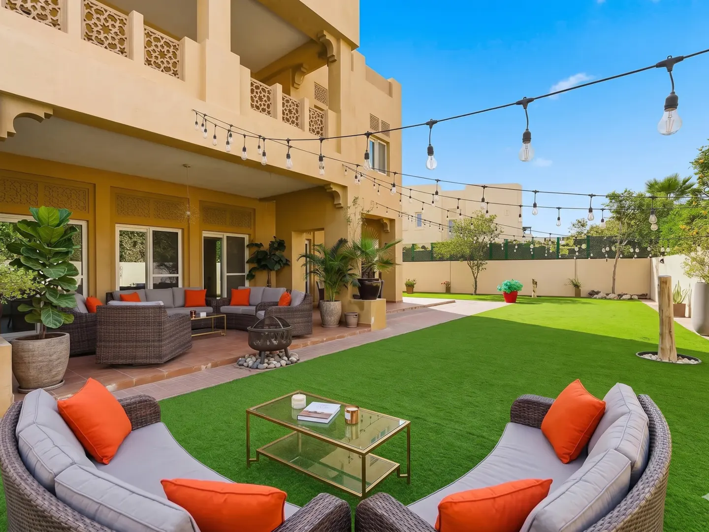 Spacious backyard with a large artificial lawn, patio seating, string lights, and a tan villa with decorative balconies.