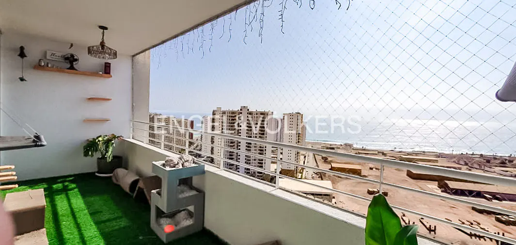 A balcony with green turf, a cat tree, and a view of the ocean and city buildings. A safety net is installed.